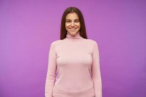 intérieur photo de Jeune longue aux cheveux brunette Dame à la recherche positivement à caméra avec charmant sourire, en gardant sa mains vers le bas tandis que permanent plus de violet Contexte