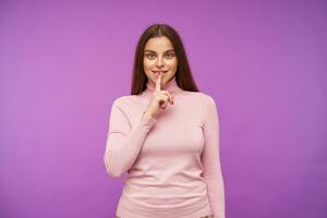 positif Jeune aux yeux verts marron aux cheveux femme habillé dans rose col roulé chandail en gardant index sur sa kips tandis que souriant agréablement à caméra, posant plus de violet Contexte photo