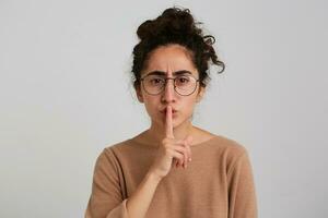 portrait de malheureux, renfrogné fille avec foncé frisé cheveux chignon. portant beige sauteur et lunettes. émotion concept. montrant silence signe, être calme. en train de regarder à le caméra isolé plus de blanc Contexte photo