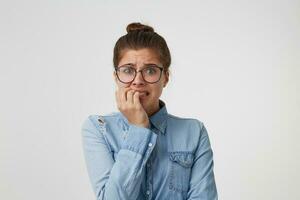 portrait de une fille étudiant dans lunettes, habillé dans une à la mode denim chemise, des soucis à propos quelque chose, nerveux, mord sa clous, attendre pour le examen résultats isolé sur blanc Contexte photo