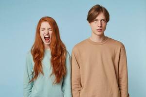 couple relation. une fille avec rouge longue cheveux des stands avec yeux fermé ouverture sa bouche large comme si en hurlant, le gars suivant à sa est roulant le sien yeux en haut fatigué de écoute à le pleure, de disputes photo