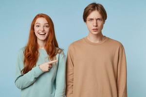 studio photo de une couple, fille dans une bien humeur, joyeuse sourires, spectacles avec une index à le gars qui des stands avec une confus, incompréhension méfiant faciale expression, isolé sur bleu Contexte.