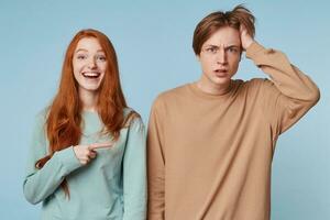 proche en haut de couple, fille avec longue rouge cheveux des rires spectacles avec sa indice doigt à le gars est permanent avec une déconcerté, perplexe expression, dans panique, agrippant le sien tête avec le sien main, bleu Contexte photo