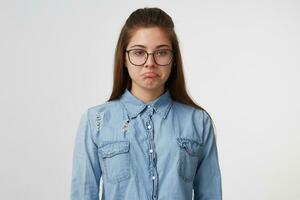 portrait de une Jeune jolie fille dans des lunettes avec longue cheveux regards triste dérangé malheureux faire la moue lèvres, habillé dans une denim chemise isolé sur blanc Contexte photo