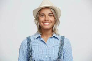 joyeux mignonne fille souriant, à la recherche à le caméra, habillé dans décontractée vêtements et une blanc chapeau, magnifique agréable amical femme, isolé plus de blanc Contexte. photo