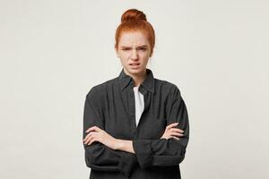 une roux fille avec sa cheveux plié dans une chignon des stands avec bras franchi se sent dégoûté, habillé dans noir Pour des hommes chemise, isolé plus de blanc fond.grimaced de quelque chose désagréable. photo