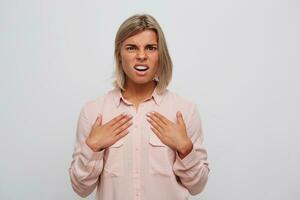 fermer de irrité mécontent blond Jeune femme avec un appareil dentaire sur les dents porte rose chemise se sent confus et points à se par mains isolé plus de blanc Contexte photo