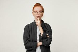 portrait de une fille avec rouge cheveux habillé dans une noir Pour des hommes chemise avec des lunettes dépeint une geste de silence en portant une doigt près le bouche, demande ne pas à faire bruit, à maintenir silence photo