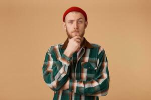 une sur de soi homme avec une barbe habillé dans cool rouge casquette et une à carreaux chemise détient le sien main sur menton, un sourcil ludique élevé pense à propos quelque chose, construire une plan, réfléchit une cool idée. photo