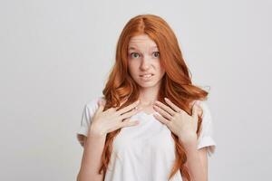 portrait de étonné mécontent roux Jeune femme avec longue cheveux et taches de rousseur porte t chemise regards étonné et montrer du doigt à se isolé plus de blanc Contexte photo