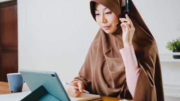 une dame musulmane d'asie porte un casque à l'aide d'une tablette numérique parle à des collègues du rapport de vente lors d'une conférence vidéo tout en travaillant à domicile dans la cuisine. distanciation sociale, quarantaine pour le virus corona. photo