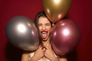 excité Jeune jolie foncé aux cheveux femme avec de fête maquillage renfrogné sa visage et en gardant bouche large ouvert tandis que permanent plus de Bourgogne Contexte avec hélium des ballons photo