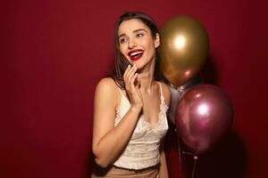 intérieur photo de Jeune jolie élégant foncé aux cheveux femelle en portant air des ballons et à la recherche positivement de côté avec agréable sourire, permanent plus de Bourgogne Contexte