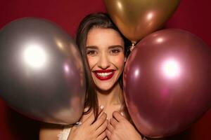 portrait de joyeux Jeune aux yeux bruns brunette Dame avec rouge lèvres posant plus de coloré air des ballons tandis que ayant agréable fête avec sa amis, étant dans haute esprit et souriant largement photo