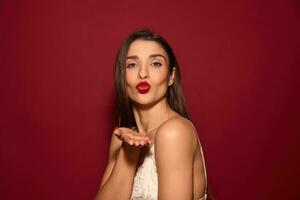 studio photo de Jeune jolie foncé aux cheveux Dame avec soir maquillage pliant sa lèvres dans baiser et en gardant sa paume élevé tandis que permanent plus de Bourgogne Contexte