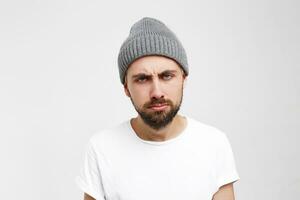 très ennuyé adulte homme avec barbe à la recherche fatigué et malade, plus de blanc Contexte. homme veut à être quelque part autre. attrayant Jeune homme dans gris chapeau, sentiment indifférent, exprimer le désir à laisser photo