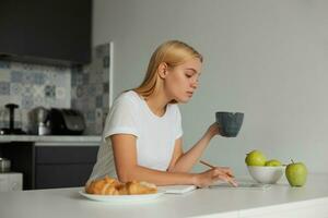 une Jeune blond fille Planification sa jour, garde une gros gris tasse, pistes le sien doigt à travers le téléphone filtrer. des croissants sur une plaque, pommes, sont là sur une blanc cuisine tableau, cuisine Contexte photo