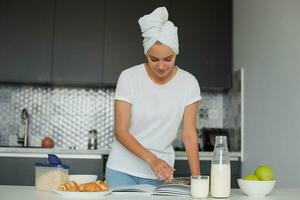 attrayant blond fille des stands près le table dans le matin, souriant, regards certains livre, Aller à avoir petit-déjeuner, habillé mine de rien et tête enveloppé dans une serviette, cuisine Contexte photo