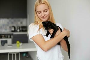 une Jeune blond fille à le cuisine détient sur mains, des caresses, caressant, câlins noir chaton, mignonne tendrement souriant, habillé dans une national blanc T-shirt, cuisine Contexte photo