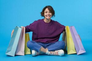 intérieur coup de heureux jolie Jeune brunette femelle avec court la Coupe de cheveux embrassant Sacs avec sa achats et souriant largement à caméra, isolé plus de bleu Contexte photo