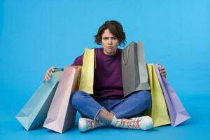 sérieux Jeune attrayant frisé brunette femme avec Naturel maquillage à la recherche avec colère à caméra et embrassement papier Sacs, séance plus de bleu Contexte dans décontractée vêtements photo