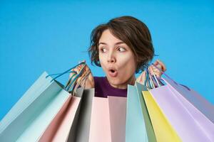 agité Jeune court aux cheveux brunette Dame avec décontractée coiffure à la recherche avec surprise à caméra avec ouvert bouche tandis que en portant papier Sacs dans élevé mains, isolé plus de bleu Contexte photo