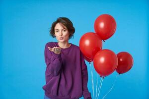 positif Jeune frisé court aux cheveux brunette Dame pliant lèvres dans air baiser et en gardant paume élevé tandis que posant plus de bleu Contexte avec bouquet de rouge air des ballons photo