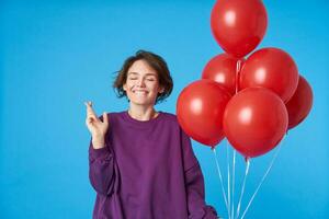 agréable à la recherche Jeune magnifique foncé aux cheveux femme avec court la Coupe de cheveux souriant volontier avec fermé yeux et traversée les doigts pour bien la chance tandis que posant plus de bleu Contexte avec rouge des ballons photo