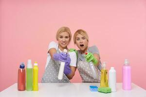 effrayé Jeune à tête blanche Dames élevage mains avec vaporisateur bouteille et à la recherche peur à caméra, étant effrayé avec Quelqu'un tandis que fabrication printemps nettoyage, isolé plus de rose Contexte photo