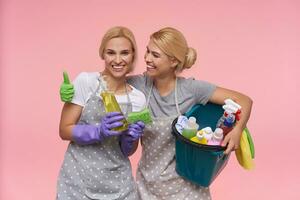 joyeux Jeune à tête blanche femmes au foyer avec chignon coiffure étant dans agréable ambiance et souriant largement tandis que permanent plus de rose Contexte dans décontractée t-shirts et polka point tabliers photo