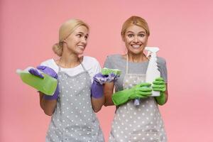 studio coup de charmant Jeune à tête blanche nettoyage Dames habillé dans uniforme tandis que nettoyage maison ensemble, permanent plus de rose Contexte avec caoutchouc gants sur leur mains photo