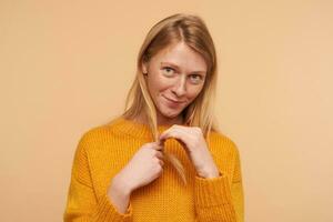 studio photo de timide aux yeux verts Jeune roux femelle tirant sa cheveux avec élevé main et à la recherche timidement à caméra, permanent contre beige Contexte