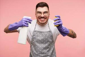 portrait de joyeux Jeune court aux cheveux brunette gars dans des lunettes Aller à laver plat et en gardant vaporisateur bouteille et éponge dans élevé mains, isolé plus de rose Contexte photo