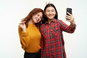 deux femmes amis. fabrication selfie sur téléphone intelligent. fille pièces avec cheveux et mettre cornes à une ami. portant Jaune chandail et à carreaux chemise. gens concept. supporter isolé plus de blanc Contexte photo