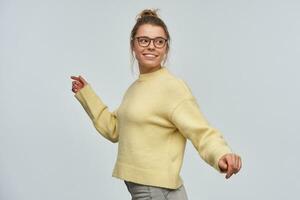 mignon, magnifique fille avec blond cheveux recueillies dans chignon. portant Jaune chandail et lunettes. en marchant une façon et en train de regarder plus de sa épaule à le droite à copie espace. supporter isolé plus de blanc Contexte photo