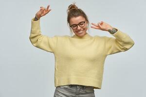 adolescent fille, content à la recherche femme avec blond cheveux recueillies dans chignon et tatouages. portant Jaune chandail et lunettes. garde sa bras levé et montrant paix panneaux. dansant isolé plus de blanc Contexte photo