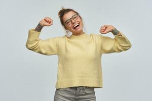 espiègle fille avec blond cheveux recueillies dans chignon et tatouages. portant Jaune chandail et lunettes. élevage sa bras, s'incline sa tête et souriant. en train de regarder à le caméra, isolé plus de blanc Contexte photo