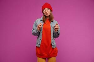 adolescent fille, content à la recherche femme avec brunette longue cheveux. portant jeans veste, Jaune pantalon, rouge chandail et chapeau. en train de regarder à le la gauche à copie espace, isolé plus de violet Contexte photo