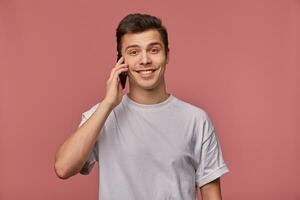 Jeune Beau de bonne humeur homme porte dans Vide T-shirt, regards à le caméra avec content expression, des stands plus de rose Contexte et parle avec ami sur le Téléphone. photo