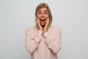 portrait de de bonne humeur excité blond Jeune femme avec un appareil dentaire sur les dents porte rose chemise émouvant sa joue, regards surpris et en criant isolé plus de blanc Contexte photo