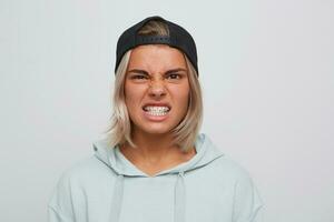 portrait de furieux en colère blond Jeune femme avec un appareil dentaire sur les dents porte noir casquette et sweat à capuche regards irrité et furieux isolé plus de blanc Contexte photo