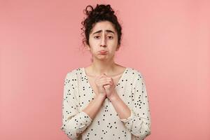 brunette fille implorant mendicité, fait la moue sa joues, regards triste chérir espoir, plié mains dans une prier geste, habillé dans chemisier avec polka des points, Aller à pleurer, isolé sur rose photo