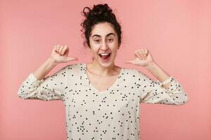 satisfait Jeune attrayant frisé brunette fille avec marron yeux dans blanc chemisier avec polka points montrer du doigt avec dé à coudre à se dans excitation étant choisi ou une gagnant dans concours, plus de rose mur photo