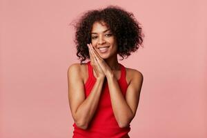 portrait de satisfait africain américain femme avec afro coiffure se sent plaisir, sourires, se réjouit, plié mains comme Aller à dormir, avoir compliments flirte, portant rouge maillot, isolé sur rose mur photo