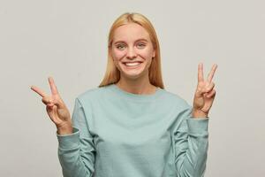 charmant Jeune blond fille a amusant, sourires, spectacles paix la victoire signe avec les doigts sur tous les deux mains, portant bleu décontractée sweat-shirt, isolé plus de gris Contexte photo