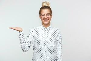 souriant content Jeune blond fille dans une bien ambiance des stands, habillé dans une blanc chemise avec noir polka des points, lunettes, cheveux recueillies dans une chignon, détient copie espace sur un palmier, sur une blanc Contexte photo