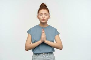 Jeune dame, jolie femme avec gingembre cheveux chignon. portant bleu T-shirt. détient sa paumes dans prière et demander, plaider pour quelque chose. émotion concept. en train de regarder à le caméra, isolé plus de blanc Contexte photo