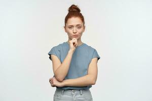 Jeune dame, méfiant à la recherche femme avec gingembre cheveux chignon. portant bleu T-shirt. émouvant sa menton et sacs à main sa lèvres. émotion concept. en train de regarder à le caméra, isolé plus de blanc Contexte photo