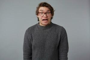 portrait de Jeune sous le choc gars avec lunettes, des stands plus de gris Contexte avec large ouvert bouche et yeux, avec surpris expression, regards étourdi. photo