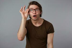 portrait de Jeune demandé homme avec des lunettes porte dans de base T-shirt, des stands plus de gris Contexte et regards à le caméra creux lunettes. photo